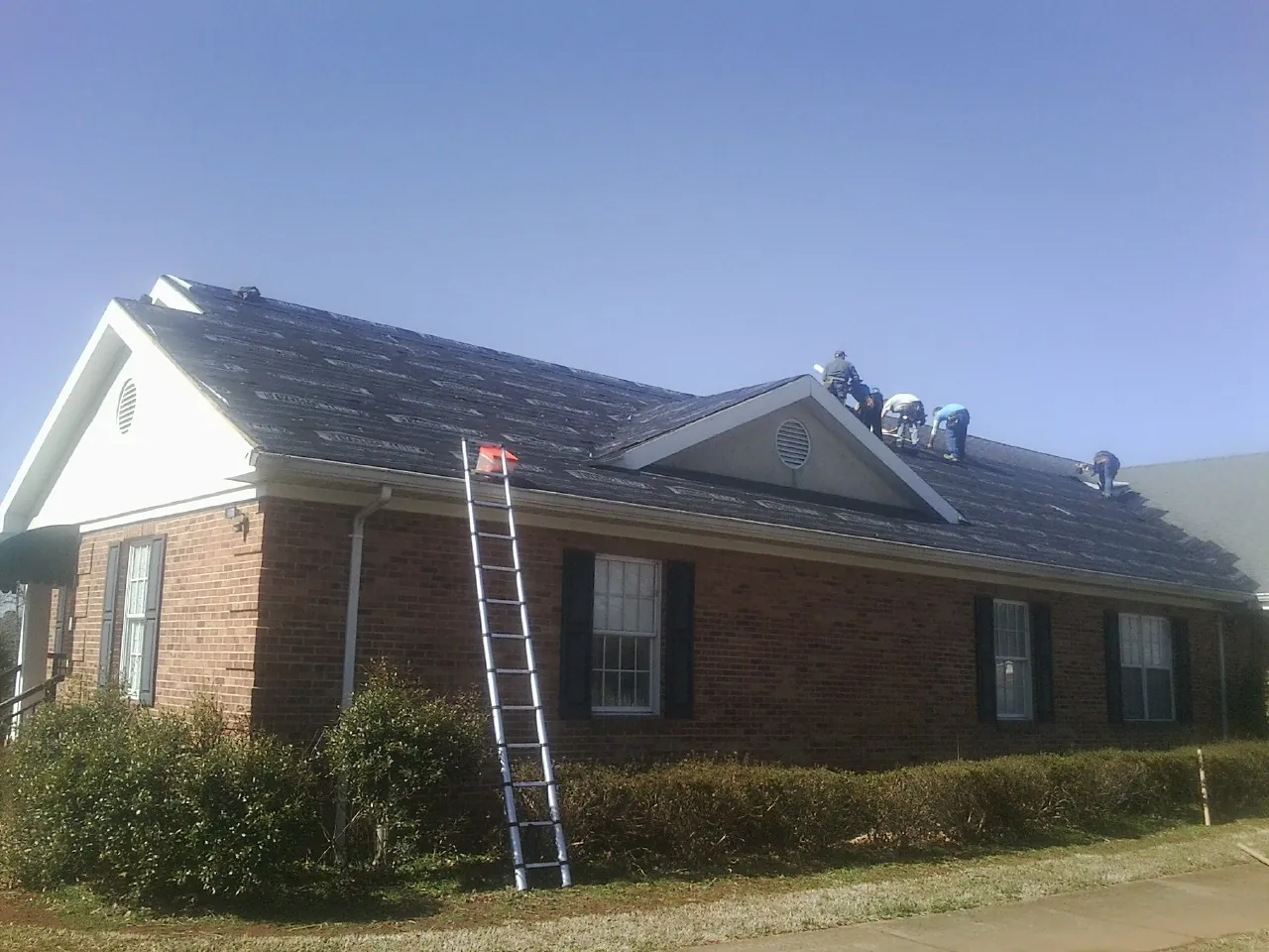 Crew tearing shingles off a brick ranch roof with a ladder set — Emergency Roof Repair work by our crew in Lima