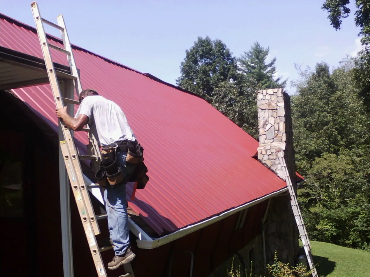 Roofer on a ladder beside a red metal roof and stone chimney — Soffit & Fascia Repair by Roofing Brothers in Lima